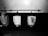 A black and white image featuring three urinals mounted on a tiled wall. The urinals are placed beneath a long horizontal bar with hooks. One urinal has a piece of paper with text attached to it.