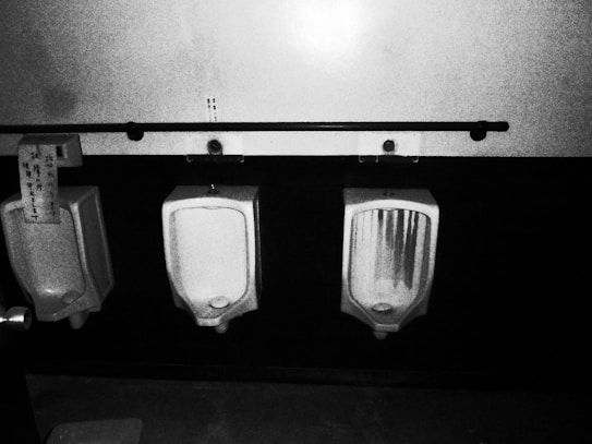 A black and white image featuring three urinals mounted on a tiled wall. The urinals are placed beneath a long horizontal bar with hooks. One urinal has a piece of paper with text attached to it.