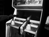A black and white photograph of a vintage arcade shooting game machine with the title 'Clay Champ' visible on the top. Two rifle-like controllers are mounted in front of the machine. The surrounding area appears dimly lit, focusing attention on the arcade machine.