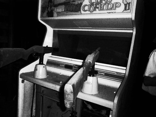 A black and white photograph of a vintage arcade shooting game machine with the title 'Clay Champ' visible on the top. Two rifle-like controllers are mounted in front of the machine. The surrounding area appears dimly lit, focusing attention on the arcade machine.