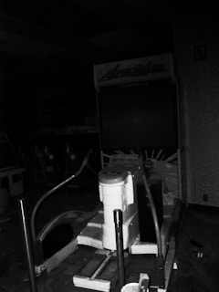 A vintage arcade game machine is situated in a dimly lit room. The console is partially visible, featuring a control panel with buttons and a seat attached to it. The surroundings are cluttered, indicating a possible storage or abandoned area.
