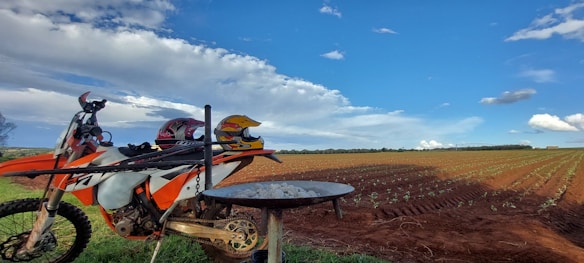 A dirt bike is parked next to a plowed agricultural field under a partially cloudy blue sky. There are two helmets placed on the bike's seat, and a charcoal grill is positioned in the foreground with white stones on it.