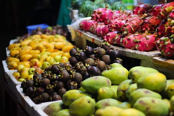 A vibrant display of assorted fruits arranged on a market stand. Bright pink dragon fruits, yellow mangoes, and dark purple mangosteens are prominently featured, creating a colorful and inviting scene. The fruits are organized into neat piles on white trays, typical of a bustling market setting.