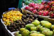 A vibrant display of assorted fruits arranged on a market stand. Bright pink dragon fruits, yellow mangoes, and dark purple mangosteens are prominently featured, creating a colorful and inviting scene. The fruits are organized into neat piles on white trays, typical of a bustling market setting.