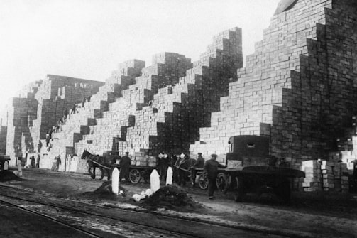 A large number of stacked stone bricks form towering structures alongside a busy working environment. Several men are involved in labor, some guiding horse-drawn carts loaded with materials, while others are engaged near the stacks. The scene depicts a historical industrial setting with early transportation methods.