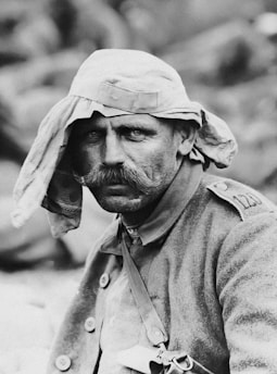 A man with a thick mustache wears a cap with fabric draped over his head for protection. He is dressed in a military-style uniform with visible buttons and straps. The expression on his face is serious, with a hint of determination. The background is blurred, focusing attention on the man's face and attire.