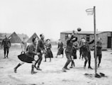 A group of soldiers in vintage uniforms are actively playing a game of netball outdoors. Several players are attempting to catch or intercept the ball near the hoop. The setting appears to be a military camp with tents and a wooden building in the background. The mood is lively and dynamic as the players are engaged in the game.