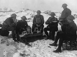 A group of six soldiers in uniform are taking a break and eating a meal together in a snowy outdoor setting. They are wearing helmets and sitting on crates and makeshift seats. Various items such as trays, cups, and containers are spread out in front of them.