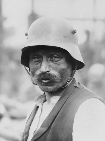 A black and white portrait of a man wearing a steel helmet with rivets visible. The man has a thick mustache and appears to be wearing a vest over a light-colored shirt. His expression is somber, and the background is blurred, focusing attention on his face.