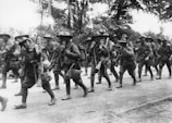 A group of soldiers in uniform are marching along a dirt road, carrying rifles and wearing wide-brimmed hats. The scene is set in a wooded area, and the soldiers seem to be moving in formation, suggesting a military operation or training exercise.