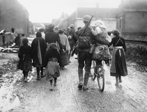 A group of people, including a soldier carrying a child on his back and pushing a bicycle, are walking down a muddy street in what appears to be a rural village setting. The individuals are dressed in early 20th-century attire, likely around wartime, suggesting an evacuation or movement of refugees.