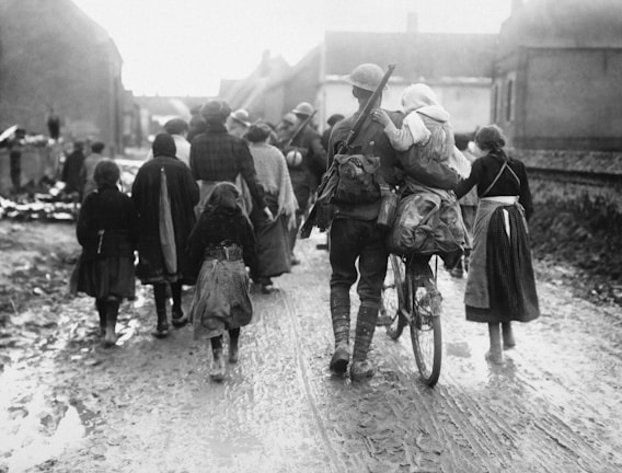 A group of people, including a soldier carrying a child on his back and pushing a bicycle, are walking down a muddy street in what appears to be a rural village setting. The individuals are dressed in early 20th-century attire, likely around wartime, suggesting an evacuation or movement of refugees.