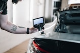A person is using a handheld device with a screen, possibly an electronic tool or microscope, to inspect the rear part of a car. The focus is on the equipment and the car’s rear section, which is slightly open, revealing some of the car's interior structure. The person's arm is visible with a tattoo and a watch.