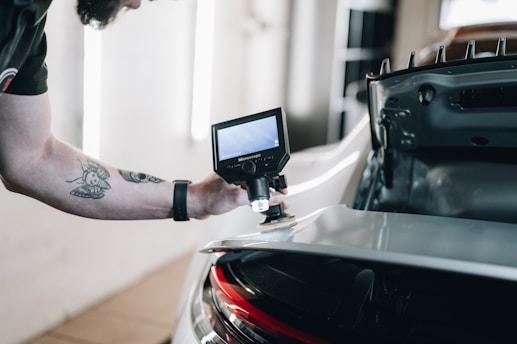 A person is using a handheld device with a screen, possibly an electronic tool or microscope, to inspect the rear part of a car. The focus is on the equipment and the car&rsquo;s rear section, which is slightly open, revealing some of the car's interior structure. The person's arm is visible with a tattoo and a watch.