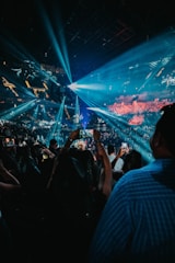 A large crowd gathers at a concert or event inside a dark arena. Bright blue and white stage lights illuminate the venue, creating beams that fan out over the audience. Many people are holding up phones, capturing the moment. The arena is filled with energy and anticipation.