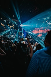 A large crowd gathers at a concert or event inside a dark arena. Bright blue and white stage lights illuminate the venue, creating beams that fan out over the audience. Many people are holding up phones, capturing the moment. The arena is filled with energy and anticipation.