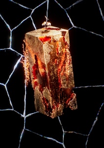 A golden rectangular object with molten, fiery textures appears suspended against a black background, crisscrossed by bright, lightning-like cracks. The central figure resembles a block of metal or stone, with glowing red and orange elements suggesting heat or energy. At the top, two small spherical objects are visible, seemingly resting on the block.