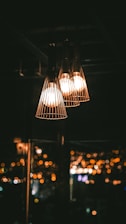 Three pendant lights hang in a dark room, casting a warm glow. The lights have a cage-like design, adding an industrial touch. In the background, city lights are blurred, creating a bokeh effect and enhancing the cozy atmosphere.
