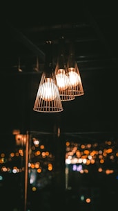 Three pendant lights hang in a dark room, casting a warm glow. The lights have a cage-like design, adding an industrial touch. In the background, city lights are blurred, creating a bokeh effect and enhancing the cozy atmosphere.