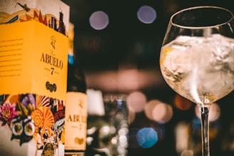 A bottle of Ron Abuelo Añejo 7 Años rum is positioned next to its vibrant packaging, which features colorful graphics including flowers and cultural symbols. In the foreground, a large glass filled with ice captures reflections and soft bokeh lights, creating a warm and inviting atmosphere.
