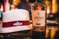 A bottle of Ron Abuelo Añejo 7 Años rum is prominently displayed on a reflective surface. Next to the bottle, there is a white hat with a maroon band, featuring the Ron Abuelo logo. The background is softly blurred, highlighting the bottle and hat in the foreground.