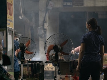 A street food vendor scene with two oscillating fans positioned in front of a stall. A person wearing a mask stands near the stall, and another individual can be seen in the background, partially obscured. The atmosphere suggests a busy market setting with a slightly smoky environment.