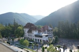A large, elegant building with a red roof and white facade surrounded by lush green trees and mountains. The building, labeled as 'Grand Cota Hotel', is situated at the base of a densely forested mountain range bathed in soft sunlight. A road with parked vehicles and neatly arranged trees runs alongside the hotel.