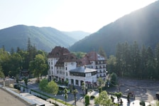 A large, elegant building with a red roof and white facade surrounded by lush green trees and mountains. The building, labeled as 'Grand Cota Hotel', is situated at the base of a densely forested mountain range bathed in soft sunlight. A road with parked vehicles and neatly arranged trees runs alongside the hotel.