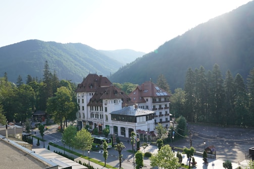A large, elegant building with a red roof and white facade surrounded by lush green trees and mountains. The building, labeled as 'Grand Cota Hotel', is situated at the base of a densely forested mountain range bathed in soft sunlight. A road with parked vehicles and neatly arranged trees runs alongside the hotel.
