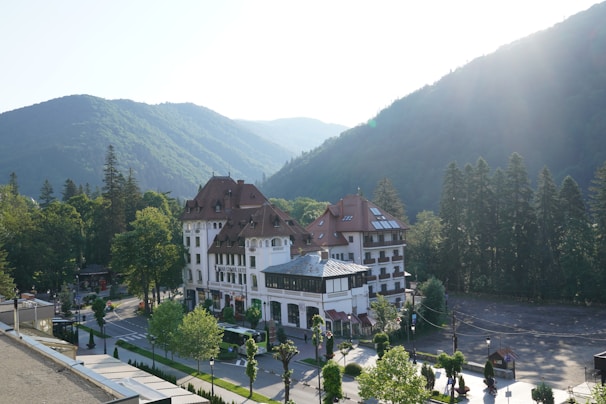 A large, elegant building with a red roof and white facade surrounded by lush green trees and mountains. The building, labeled as 'Grand Cota Hotel', is situated at the base of a densely forested mountain range bathed in soft sunlight. A road with parked vehicles and neatly arranged trees runs alongside the hotel.