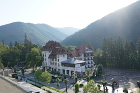 A large, elegant building with a red roof and white facade surrounded by lush green trees and mountains. The building, labeled as 'Grand Cota Hotel', is situated at the base of a densely forested mountain range bathed in soft sunlight. A road with parked vehicles and neatly arranged trees runs alongside the hotel.
