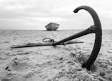 An anchor is half-buried in the sand on a beach. In the background, a small boat is blurred and tethered to the anchor. The scene is depicted in black and white, creating a timeless and serene atmosphere.