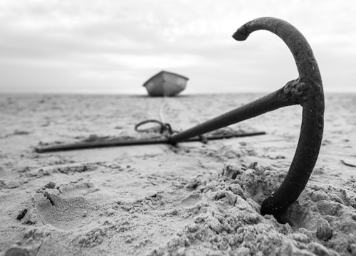 An anchor is half-buried in the sand on a beach. In the background, a small boat is blurred and tethered to the anchor. The scene is depicted in black and white, creating a timeless and serene atmosphere.