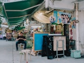 A street market scene with a mix of outdoor seating and various items for sale. A person is seated on a small stool, looking at their phone, surrounded by stacked crates, folding tables, and chairs. Above, green awnings cover the market, and posters with smiling faces are attached to the walls. The area appears somewhat cluttered with boxes and goods, suggesting an active and bustling market environment.