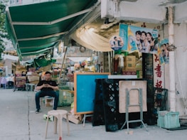 A street market scene with a mix of outdoor seating and various items for sale. A person is seated on a small stool, looking at their phone, surrounded by stacked crates, folding tables, and chairs. Above, green awnings cover the market, and posters with smiling faces are attached to the walls. The area appears somewhat cluttered with boxes and goods, suggesting an active and bustling market environment.