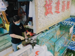 A person wearing gloves and a face mask is preparing seafood next to water tanks filled with live fish. The area is well-stocked with equipment and supplies, and there are signs with Chinese characters on the wall. The ambient lighting suggests a market or fish shop setting.