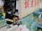 A person wearing gloves and a face mask is preparing seafood next to water tanks filled with live fish. The area is well-stocked with equipment and supplies, and there are signs with Chinese characters on the wall. The ambient lighting suggests a market or fish shop setting.