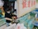A person wearing gloves and a face mask is preparing seafood next to water tanks filled with live fish. The area is well-stocked with equipment and supplies, and there are signs with Chinese characters on the wall. The ambient lighting suggests a market or fish shop setting.