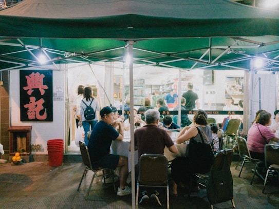 Outdoor dining area under a green canopy with several people sitting at tables. A well-lit restaurant interior is visible in the background, with more people inside. A large sign with red characters is prominently displayed on the wall next to the entrance.