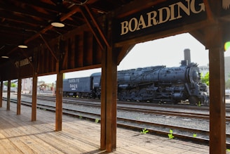 A vintage steam locomotive, labeled Santa Fe 5021, is parked on railway tracks. The scene is viewed from a wooden platform with a roof, and the word 'BOARDING' is prominently displayed on the structure. The background includes some buildings and greenery.