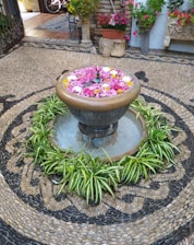 A decorative fountain is surrounded by vibrant green plants and colorful floating flowers. The area is intricately paved with a pattern of stones, providing a textured and artistic backdrop. In the background, there are potted plants with red and pink blossoms and some antique stone sculptures.