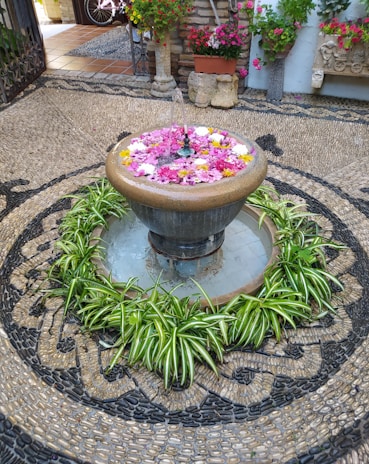 A decorative fountain is surrounded by vibrant green plants and colorful floating flowers. The area is intricately paved with a pattern of stones, providing a textured and artistic backdrop. In the background, there are potted plants with red and pink blossoms and some antique stone sculptures.