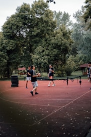 Four people are playing a casual game on an outdoor court surrounded by lush greenery. They seem engaged, with one person holding a ball. A makeshift net is set up across the court, resembling a game of volleyball or footvolley.
