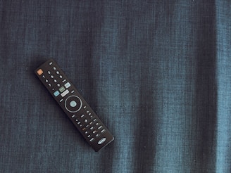 A black remote control is placed diagonally on a dark textured fabric surface. The remote has several buttons, featuring a prominent circular button in the center and a distinct orange power button.