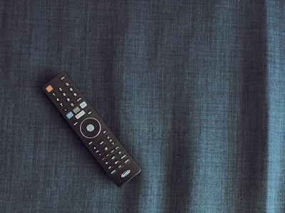A black remote control is placed diagonally on a dark textured fabric surface. The remote has several buttons, featuring a prominent circular button in the center and a distinct orange power button.