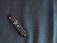 A black remote control is placed diagonally on a dark textured fabric surface. The remote has several buttons, featuring a prominent circular button in the center and a distinct orange power button.