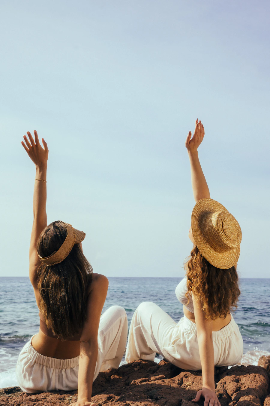 Two women in sun hats raising their arms beside the sea