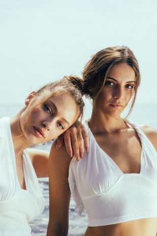 Two women are standing close together with one resting her head on the other's shoulder. Both are wearing white sleeveless tops and are in an outdoor setting, likely near the ocean as the water is visible in the background. Their expressions are calm and slightly introspective.