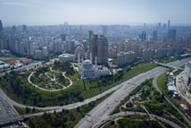 Aerial view of a large cityscape featuring a prominent white mosque with blue domes surrounded by a lush green park. High-rise buildings densely populate the background, and multiple highways with vehicles intersect throughout the scene.