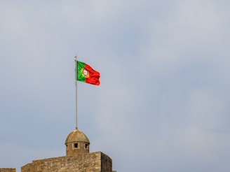 A Portuguese flag is flying atop a stone structure with a dome-shaped lookout. The sky in the background is overcast with light clouds.